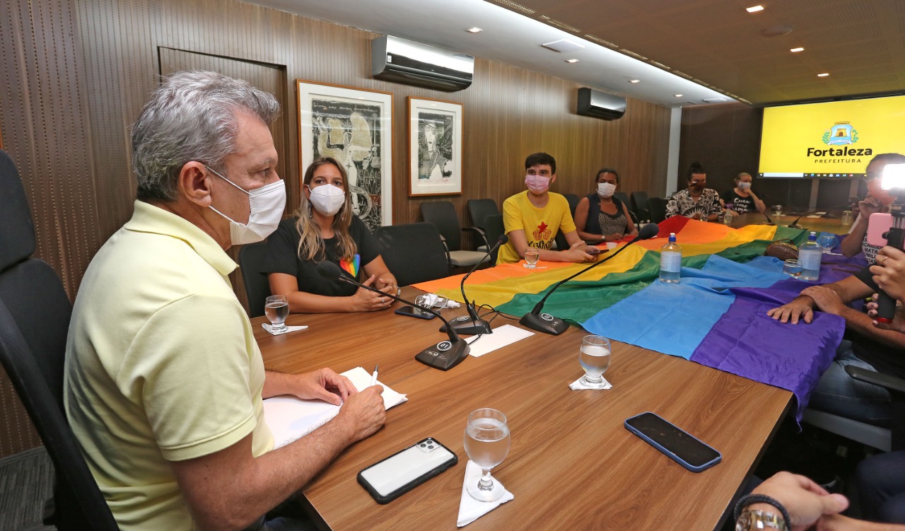 a foto mostra um grupo de pessoas em uma mesa de reunião. sobre a mesa há uma bandeira com as cores do movimento lgbt